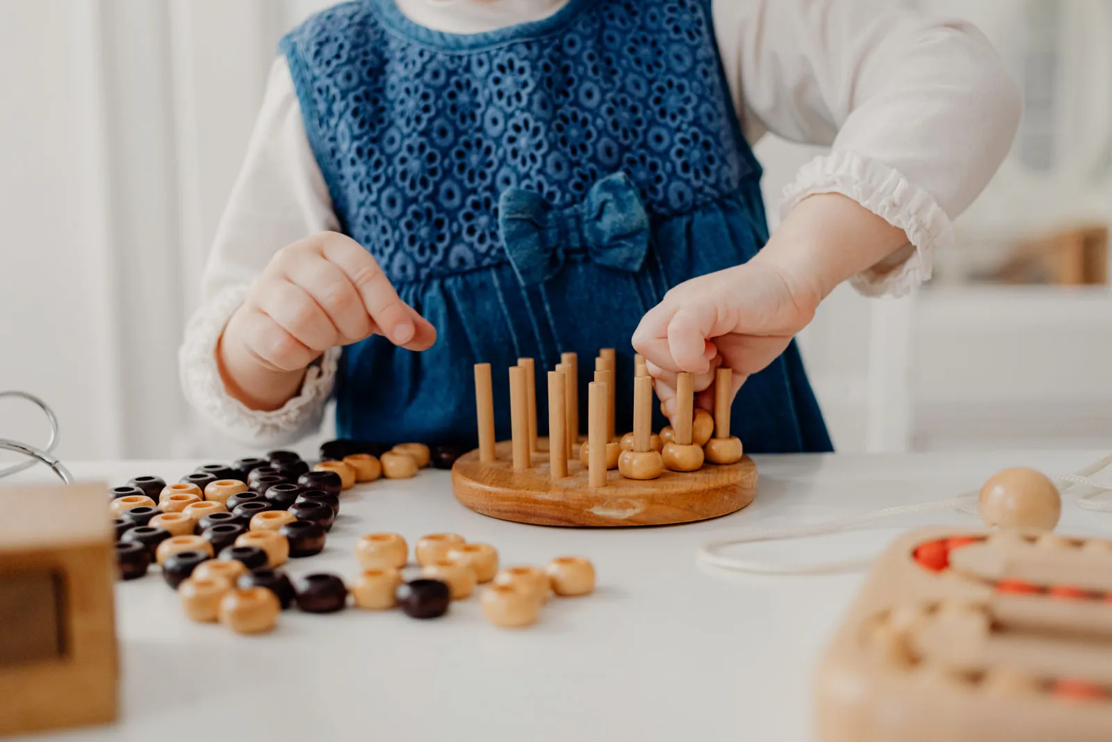 A child’s hands placing small wooden pegs into a round wooden board, with loose beads sorted across the table.