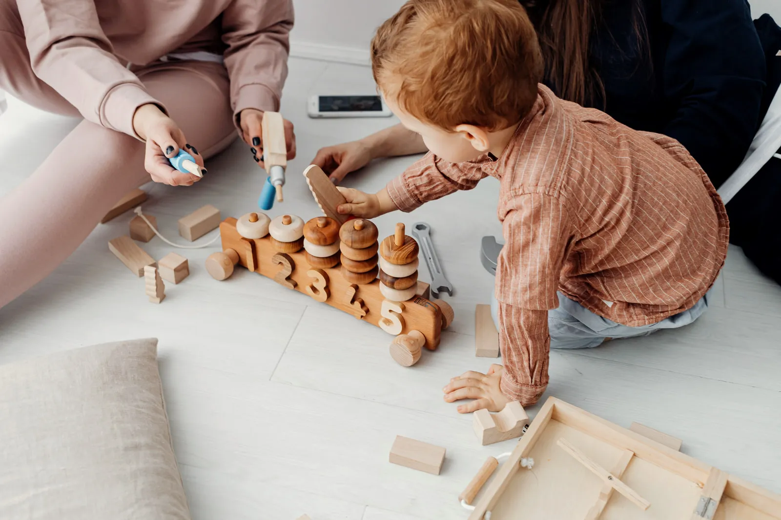 A young child kneeling on a living-room floor stacking wooden rings onto a peg, with an adult’s hands guiding from the side.