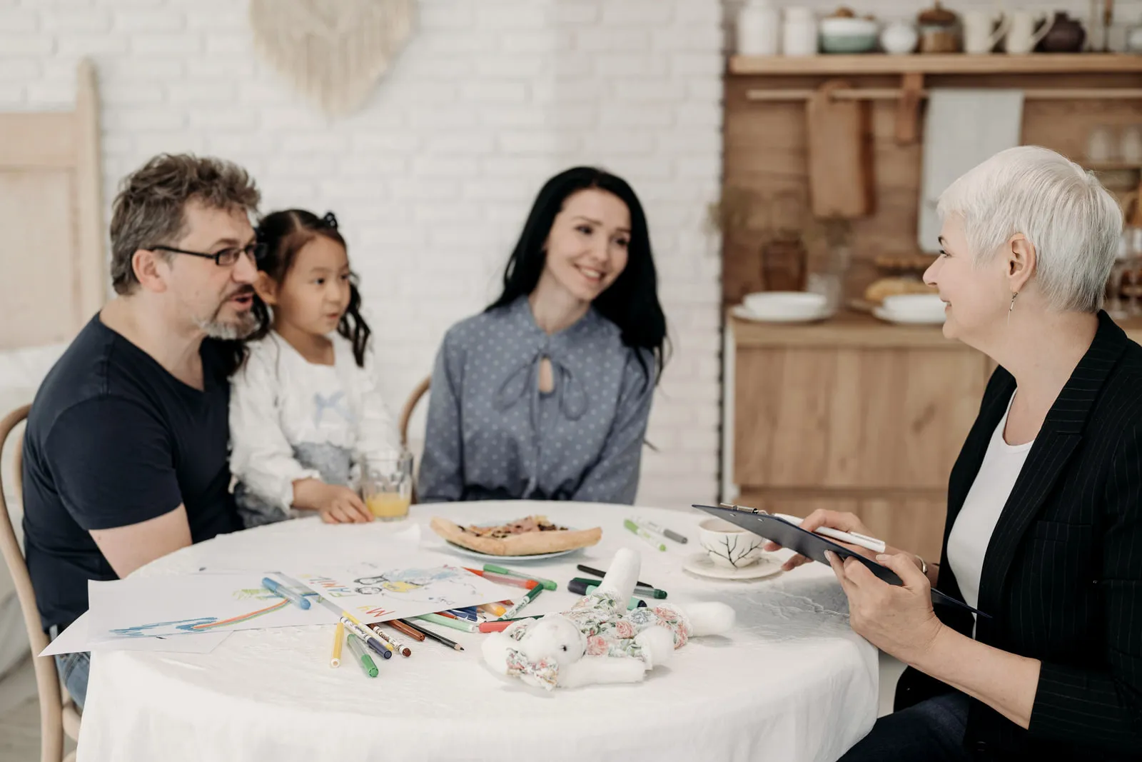 Two parents and a young child at a table with a clinician holding a tablet, talking across a spread of activity materials.