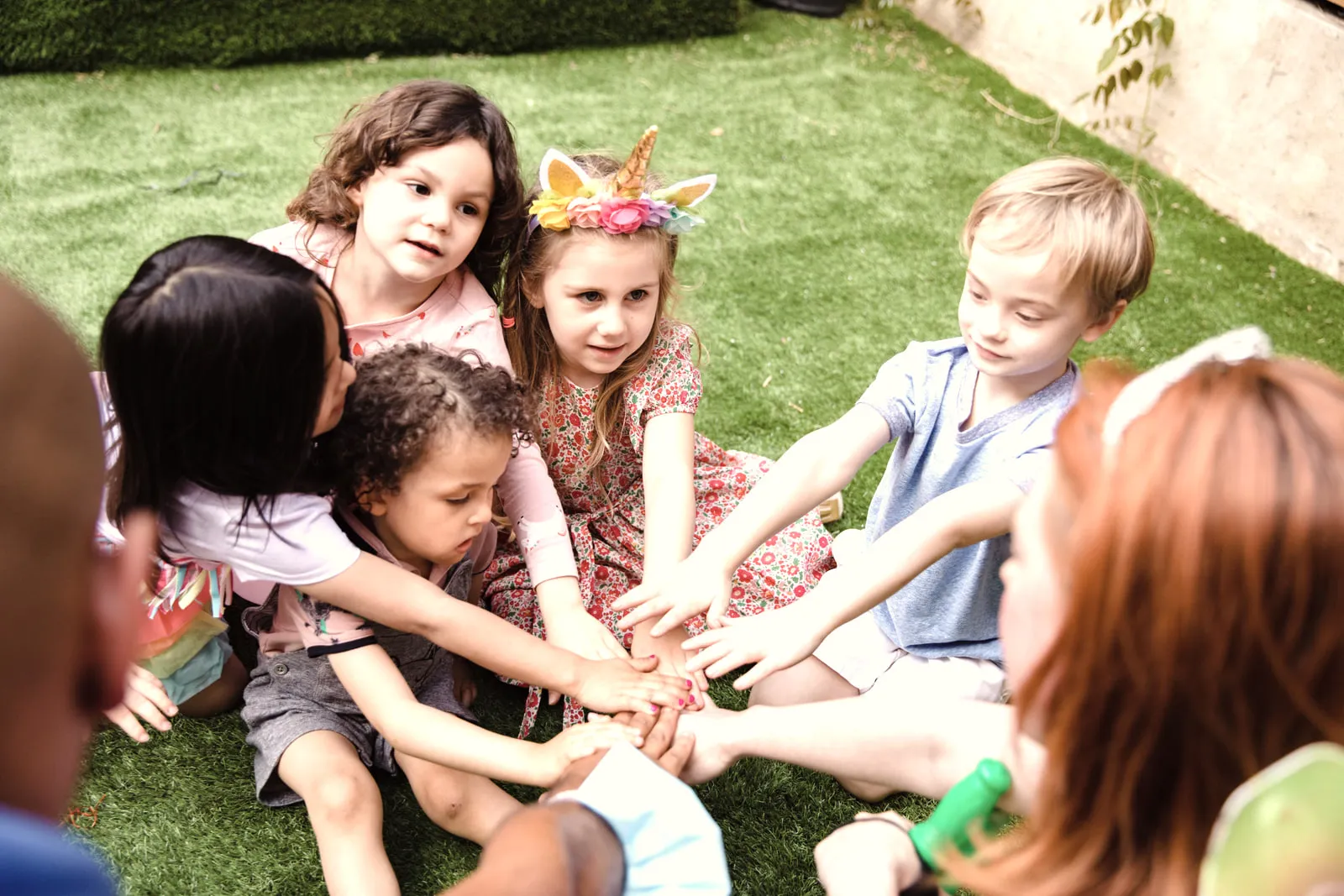 Five young children and an adult sitting in a circle on grass, all reaching their hands into the middle together.