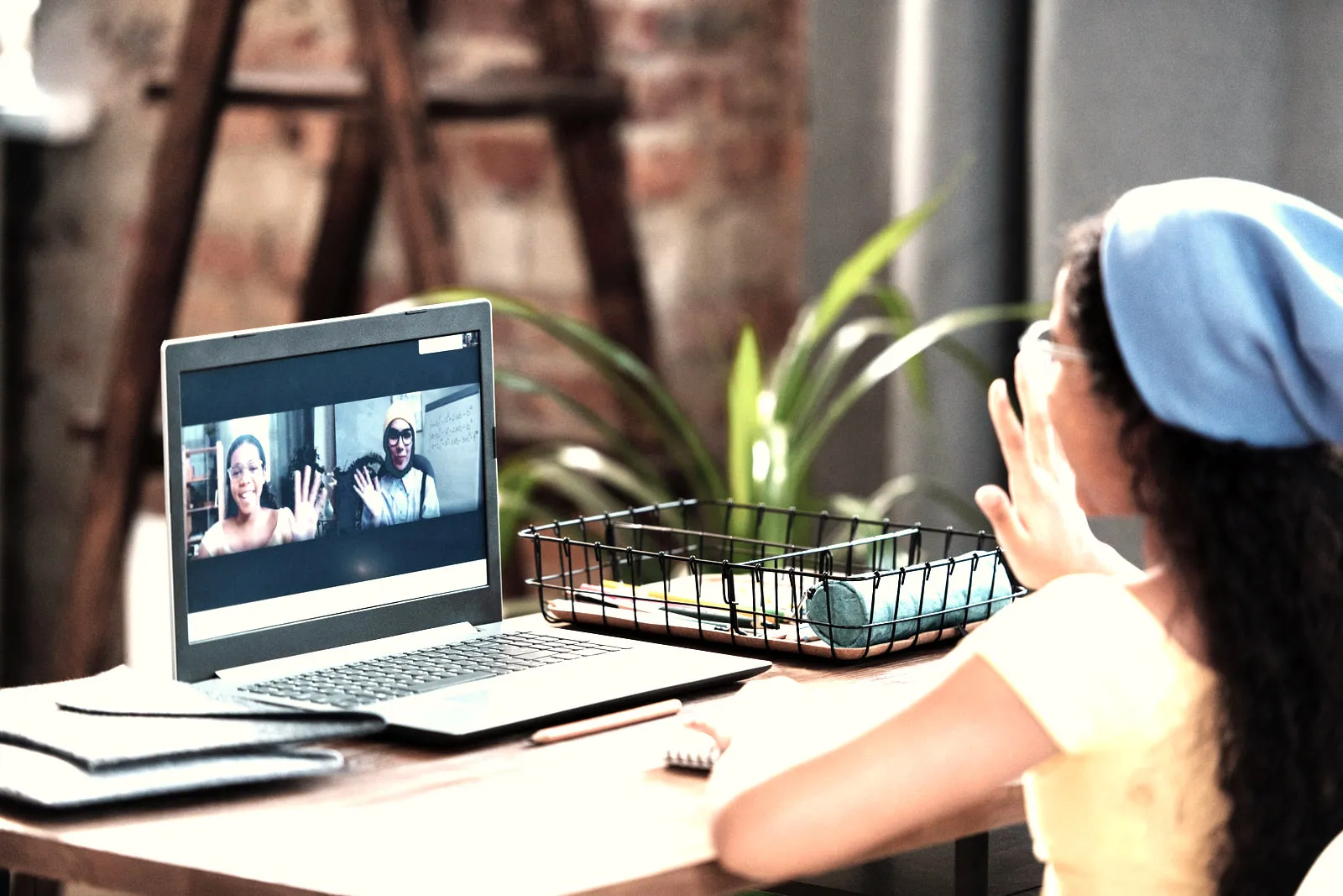 A woman at a desk raising a hand to a laptop screen showing a video call with two people on it.